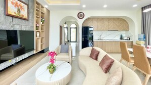 A modern living room with a sofa, coffee table, and TV. There is a kitchen area with a water dispenser and a clock on the wall. - Happy Stay Resort (Ho Chi Minh City)