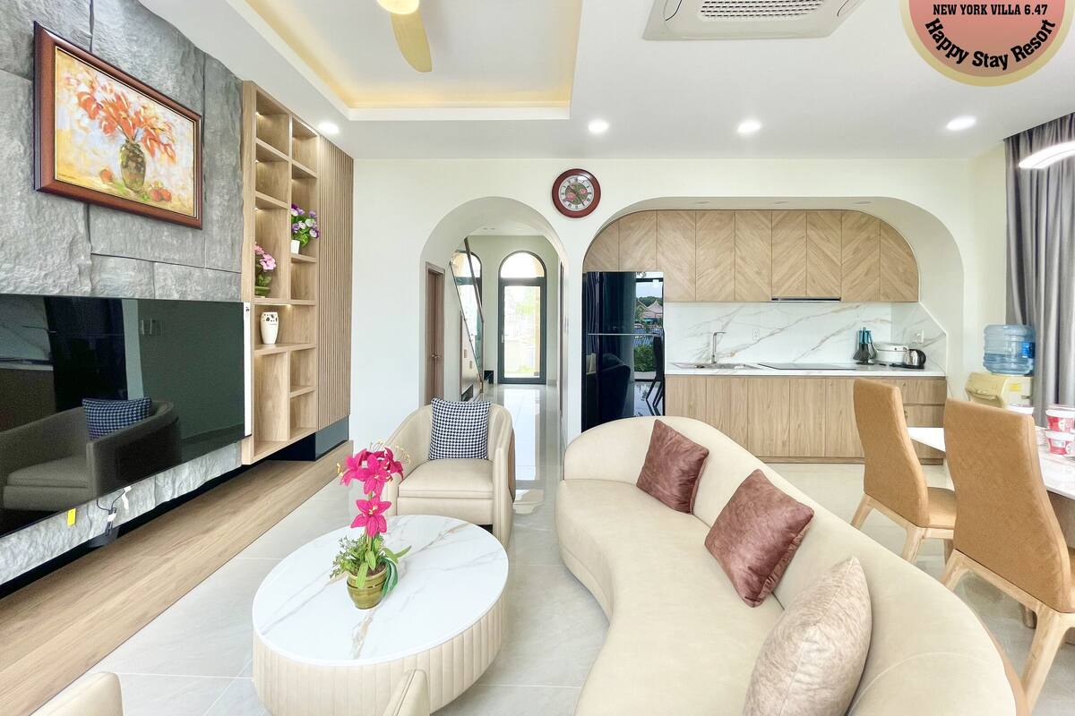 A modern living room with a sofa, coffee table, and TV. There is a kitchen area with a water dispenser and a clock on the wall.