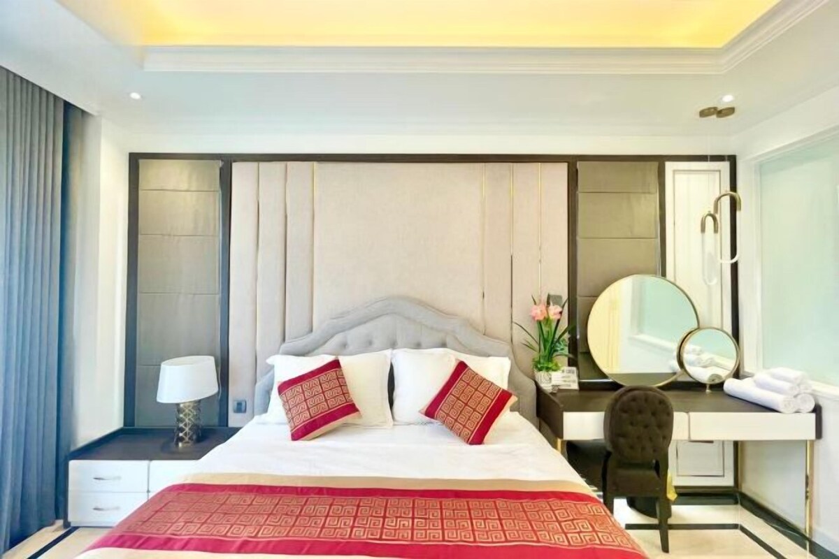 Una habitación de hotel con una cama grande, una mesita de noche con lámpara, un tocador con espejo y una silla.