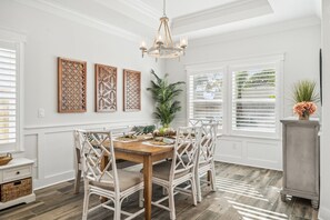 A dining area with a wooden table and white wicker chairs, surrounded by white walls and a large window with blinds. - Sunset Haven by the Pool (Panama City Beach)