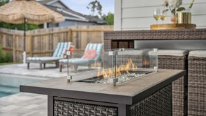 An outdoor seating area with a glass-top table, a fire feature, wicker furniture, and a pool in the background. - Sunset Haven by the Pool (Panama City Beach)