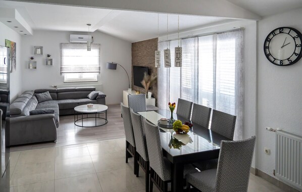 A modern living room with a grey sofa, a round coffee table, and a dining area with a glass table and grey chairs. - Luxurious home in Drum (Drum)