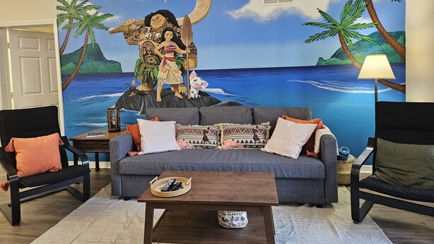 Un salon avec une grande fresque représentant un paysage tropical, un canapé gris et deux fauteuils.