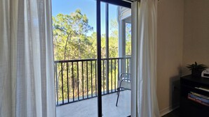 Une pièce dotée d’une grande porte-fenêtre coulissante qui donne sur un balcon agrémenté d’une rambarde en métal. On y trouve une chaise, ainsi qu’une petite table sur laquelle sont posés une plante et une horloge, sur le côté droit de la pièce.