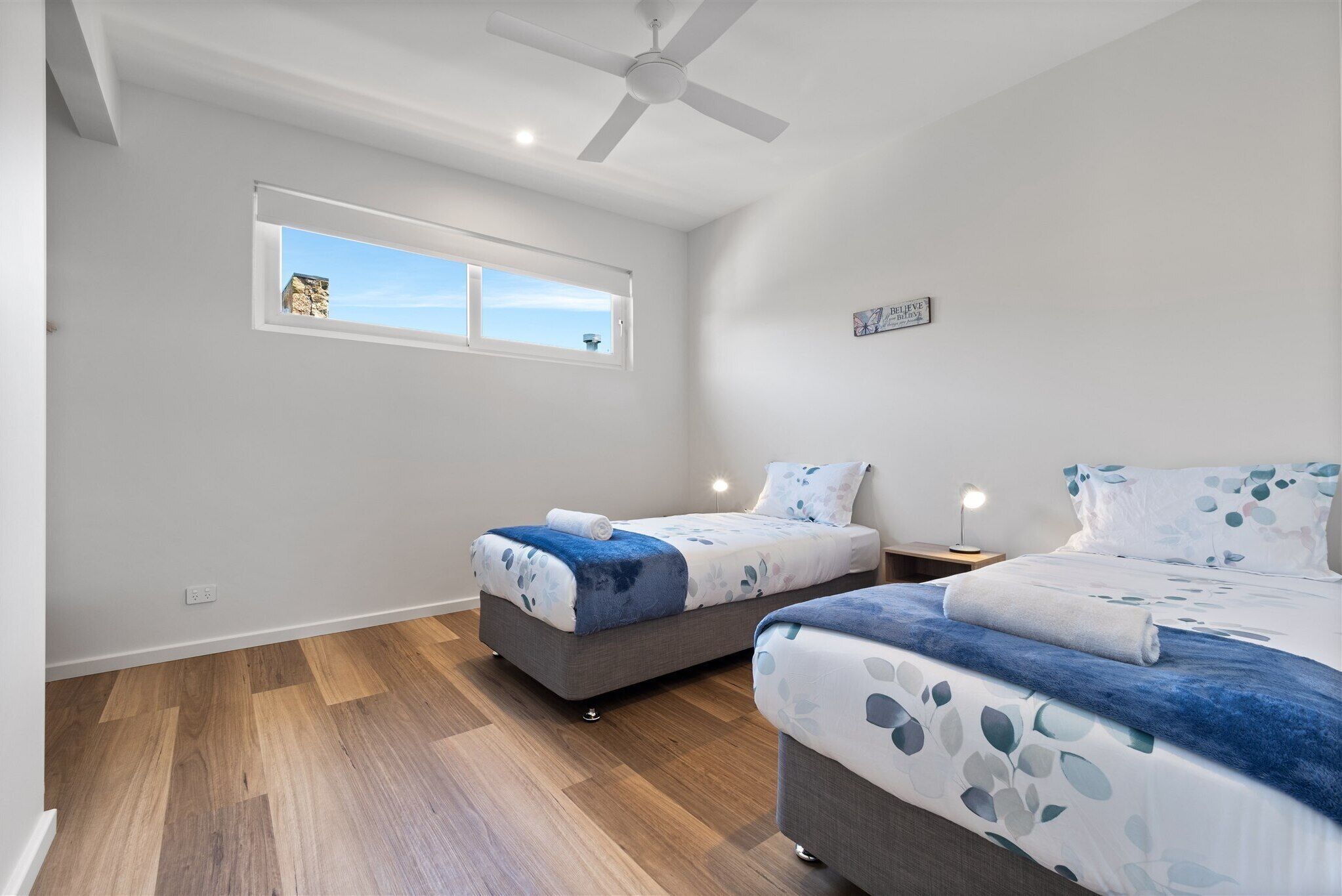 Un dormitorio con dos camas, un ventilador de techo, una ventana y una lámpara fijada a la pared.