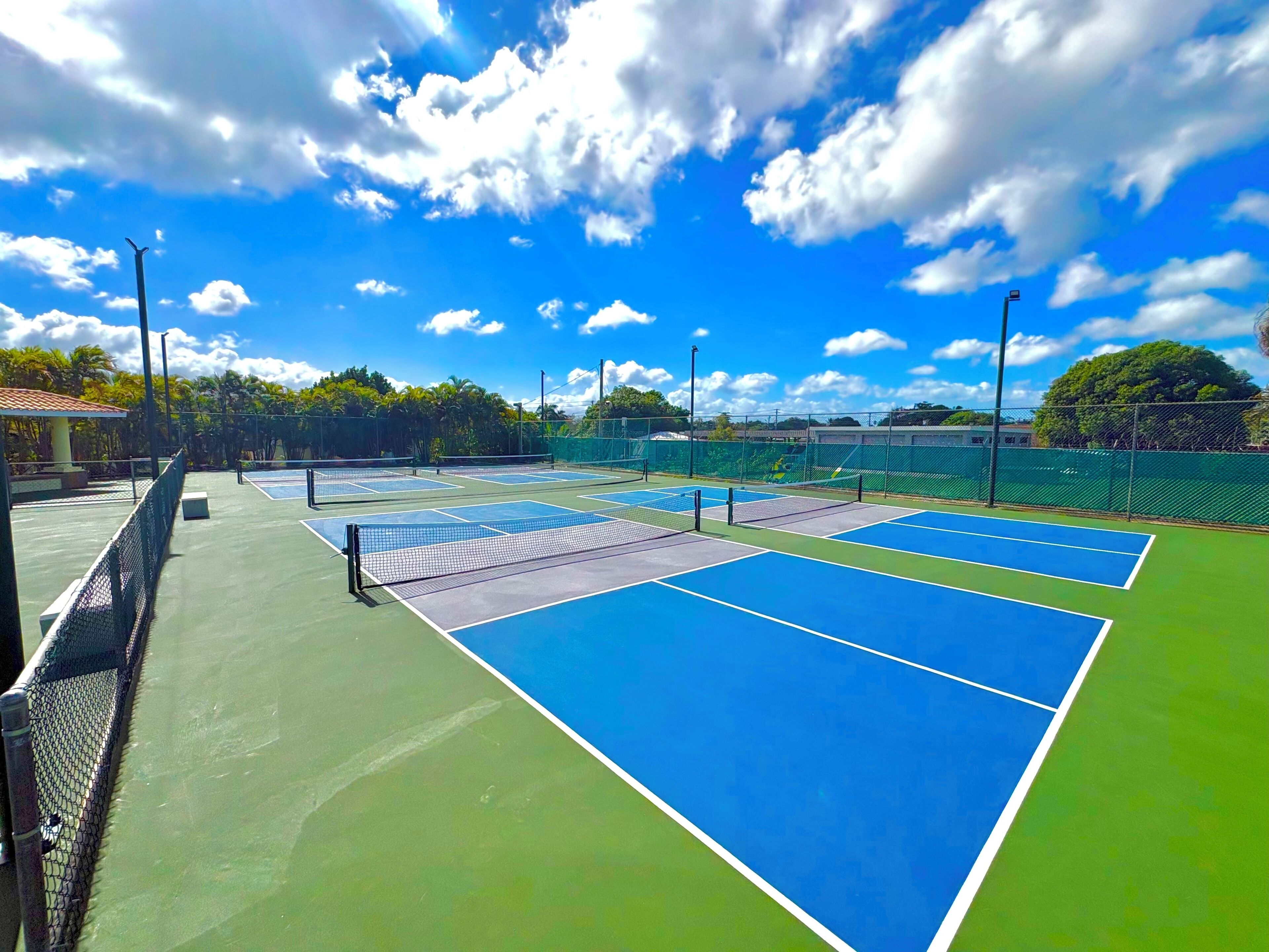 An outdoor tennis court with multiple courts, surrounded by a green fence and trees.