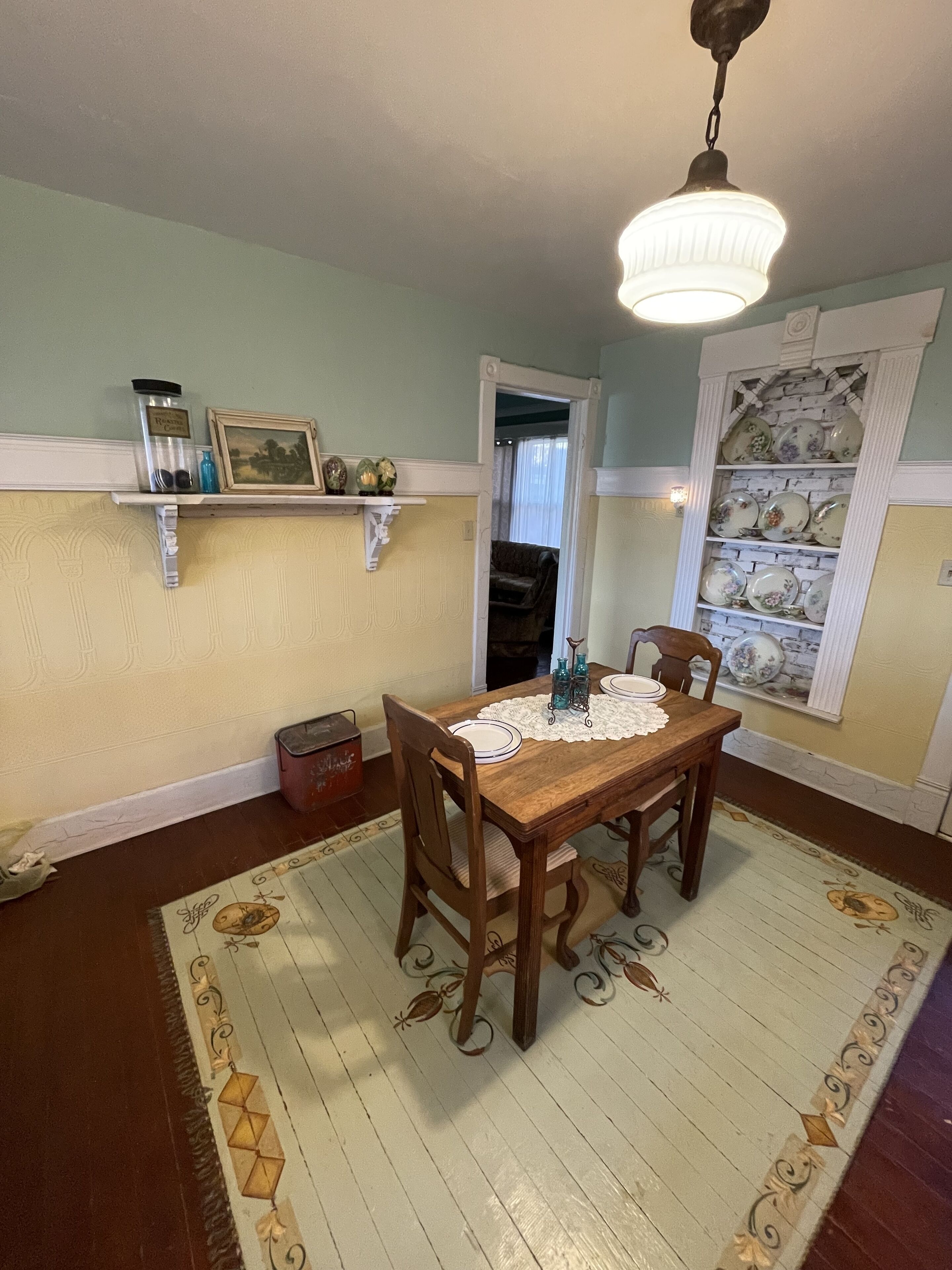 Comedor con una mesa de madera, sillas, mantel de encaje y una alfombra estampada. Hay repisas sobre la mesa con objetos decorativos y una ventana con vista al exterior.