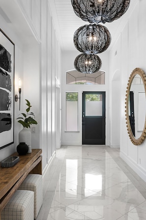 Um corredor com piso de mármore polido, um banco de madeira, uma planta em vaso, um espelho grande e um lustre singular.