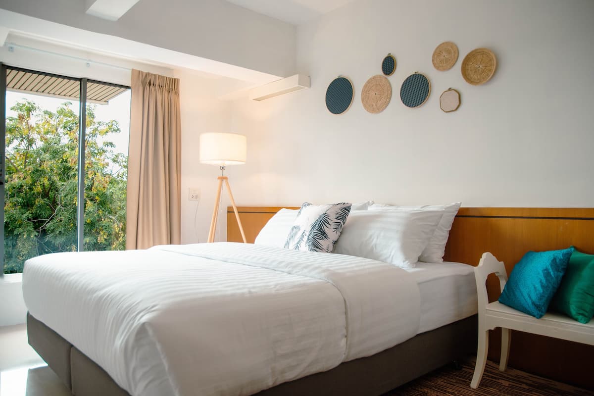 A hotel room with a large bed, a bedside table, a lamp, and a view of trees through a window.
