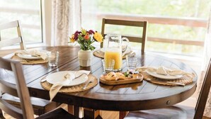 A round wooden dining table set for two with plates, cutlery, a pitcher of orange juice, a small vase with flowers, and a tray with pastries.