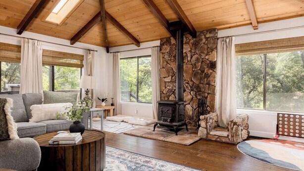 A cozy living room with a stone fireplace, wooden beams, and large windows offering a view of the outdoors.