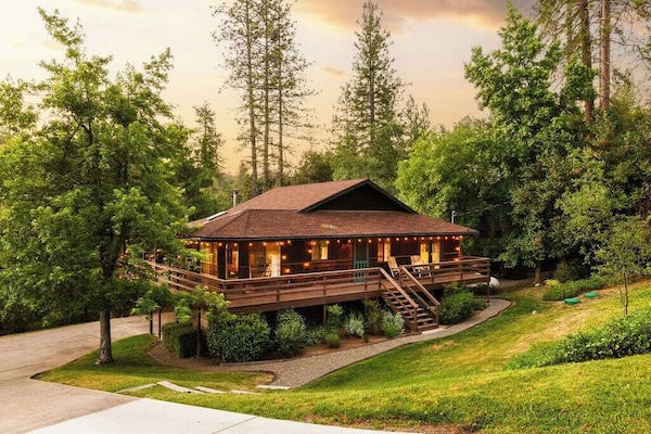Welcome to Buckeye Treehouse. A cozy wooden cabin with a wraparound deck, nestled among tall pine trees. String lights adorn the deck, creating a warm ambiance. The cabin is surrounded by lush greenery, making it a serene, natural retreat.
