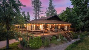 A wooden cabin with a deck, surrounded by trees and a gravel path.