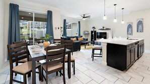 Eine moderne Küche mit Kochinsel, Essbereich mit Holzsesseln und Tisch sowie Wohnbereich mit Sofa und Fernseher.
