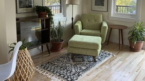 A cozy living room with a green armchair, a black and white rug, wooden flooring, and two large windows with a view of trees. - Old Town Oasis - the perfect 30 day stay (Fort Collins)