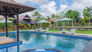 A swimming pool with lounge chairs and umbrellas, surrounded by buildings with blue roofs and greenery. - Green orry inn cafe & cottages  (Tetebatu)