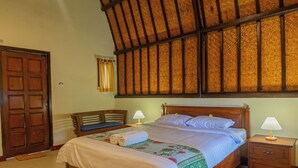 A neatly made bed with a floral bedspread and a rolled towel, a wooden nightstand with a lamp, and a wooden door. - Green orry inn cafe & cottages  (Tetebatu)