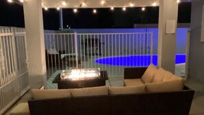 Un área de estar al aire libre con una chimenea, rodeada por una valla blanca y luces colgantes.