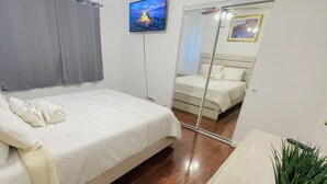 Un dormitorio con cama, espejo, televisor y planta.