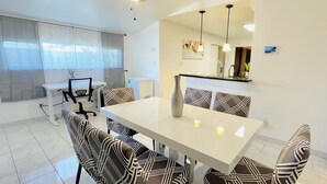 Un comedor moderno con una mesa blanca y sillas estampadas, un ventilador de techo y una zona de cocina con refrigerador y lavamanos.