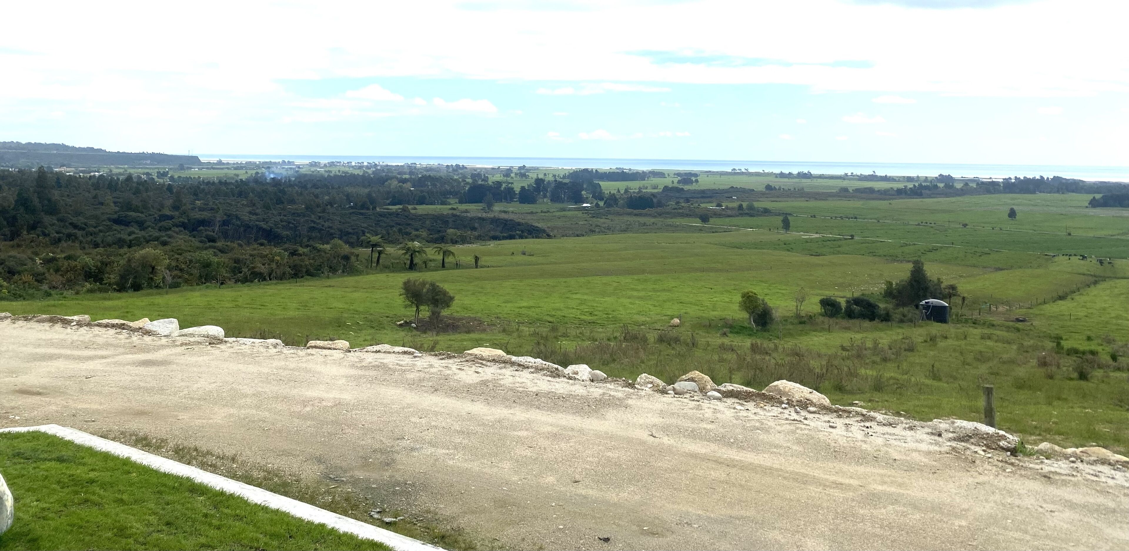 A rural landscape with a dirt road, green fields, scattered trees, and a distant view of a body of water.
