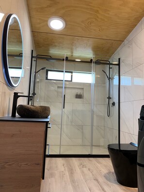 Une salle de bain moderne avec un meuble-lavabo en bois, un miroir rond, une cabine de douche en verre et une poubelle noire.