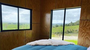 A bedroom with a large bed, wooden walls, and a view of a green landscape through two windows.