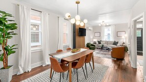 En moderne stue med et træspisebord, orange stole, en grøn sofa og et fjernsyn monteret på væggen.
