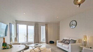 A modern living room with a white sofa, a dining table with chairs, a large window with curtains, and a clock on the wall. - West Lodge, Bognor Regis -  a flat that sleeps 4 guests  in 2 bedrooms (Bognor Regis)