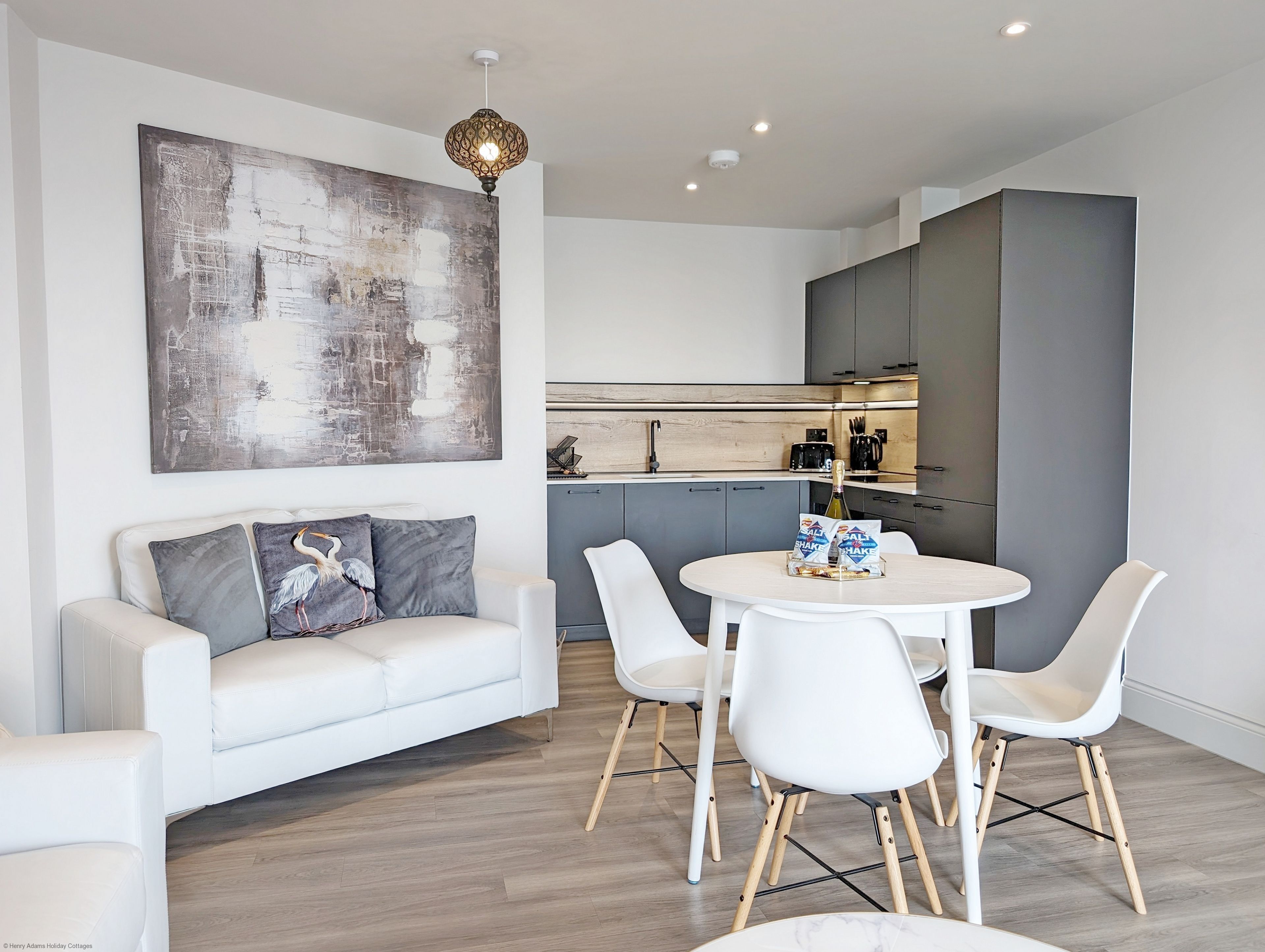 Un salon moderne avec un canapé blanc, des coussins gris, un grand tableau abstrait et un coin repas comprenant une table ronde et des chaises blanches.