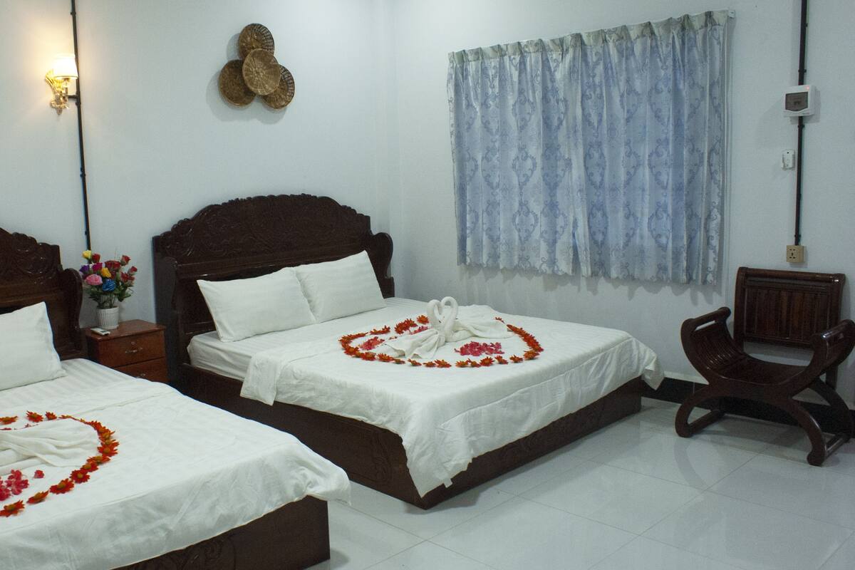 A hotel room with two beds, a nightstand, a chair, a wall-mounted light, a decorative wall piece, and a window with curtains.