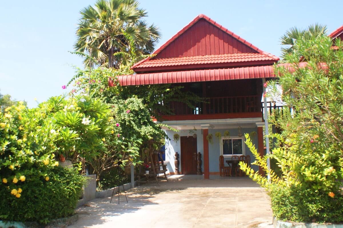 A two-story building with a red roof and a balcony. There are potted plants and a pathway leading to the entrance.