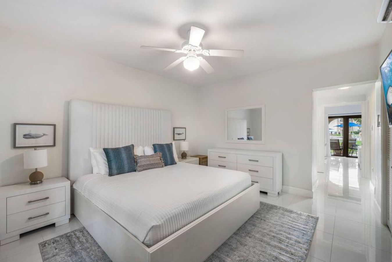 A modern bedroom with a large bed, a ceiling fan, a nightstand with a lamp, a dresser, and a mirror.