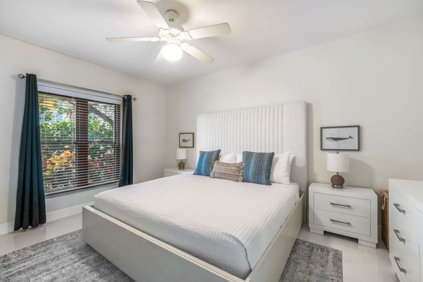 A bedroom with a bed, a nightstand, a dresser, a ceiling fan, and a window with blinds.