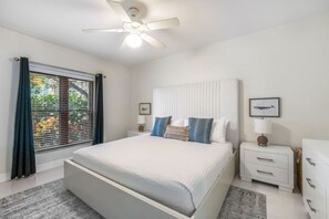 Un dormitorio con cama, mesita de noche, cómoda, ventilador de techo y ventana con persianas.