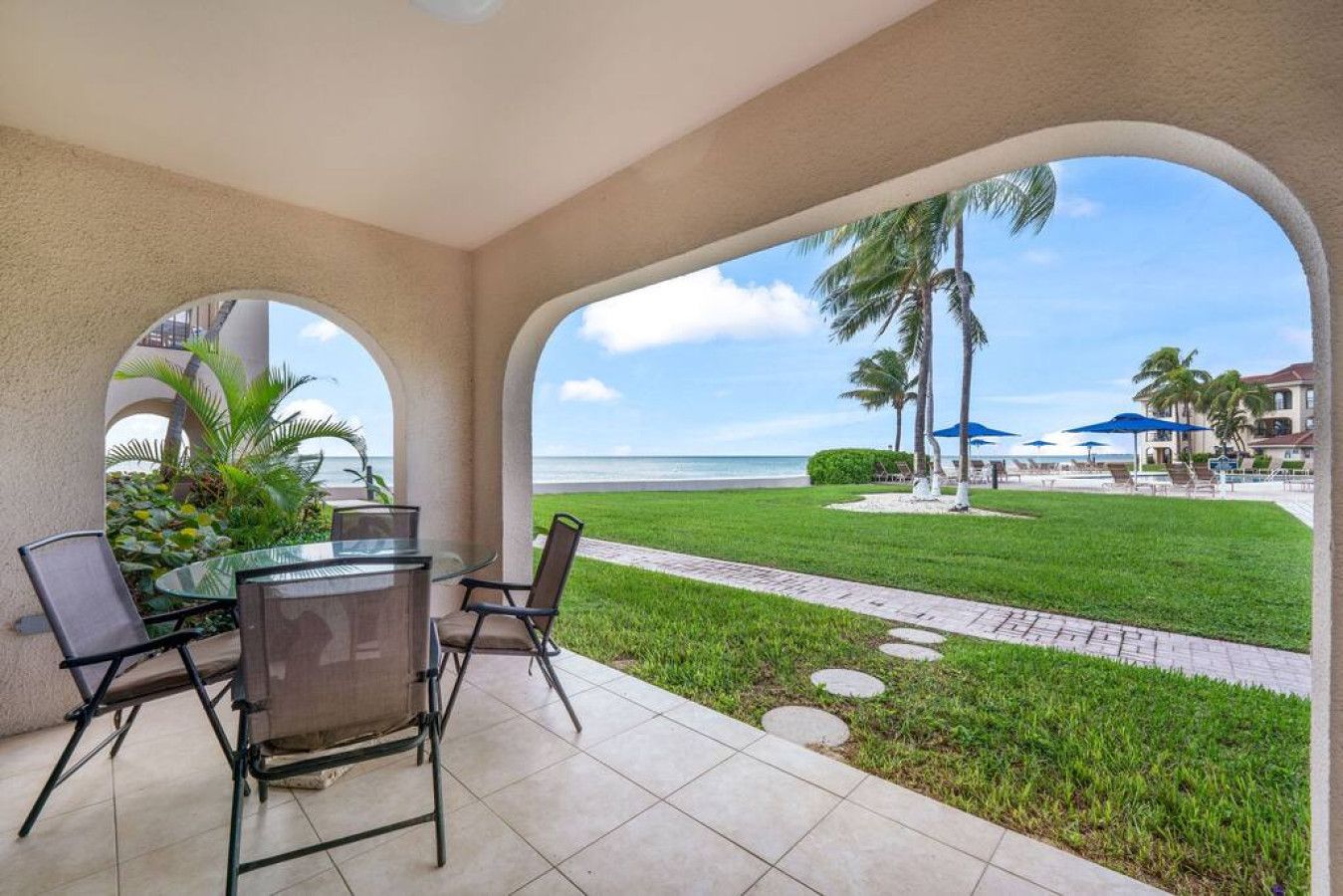 A covered outdoor seating area with a view of the beach, palm trees, and umbrellas.