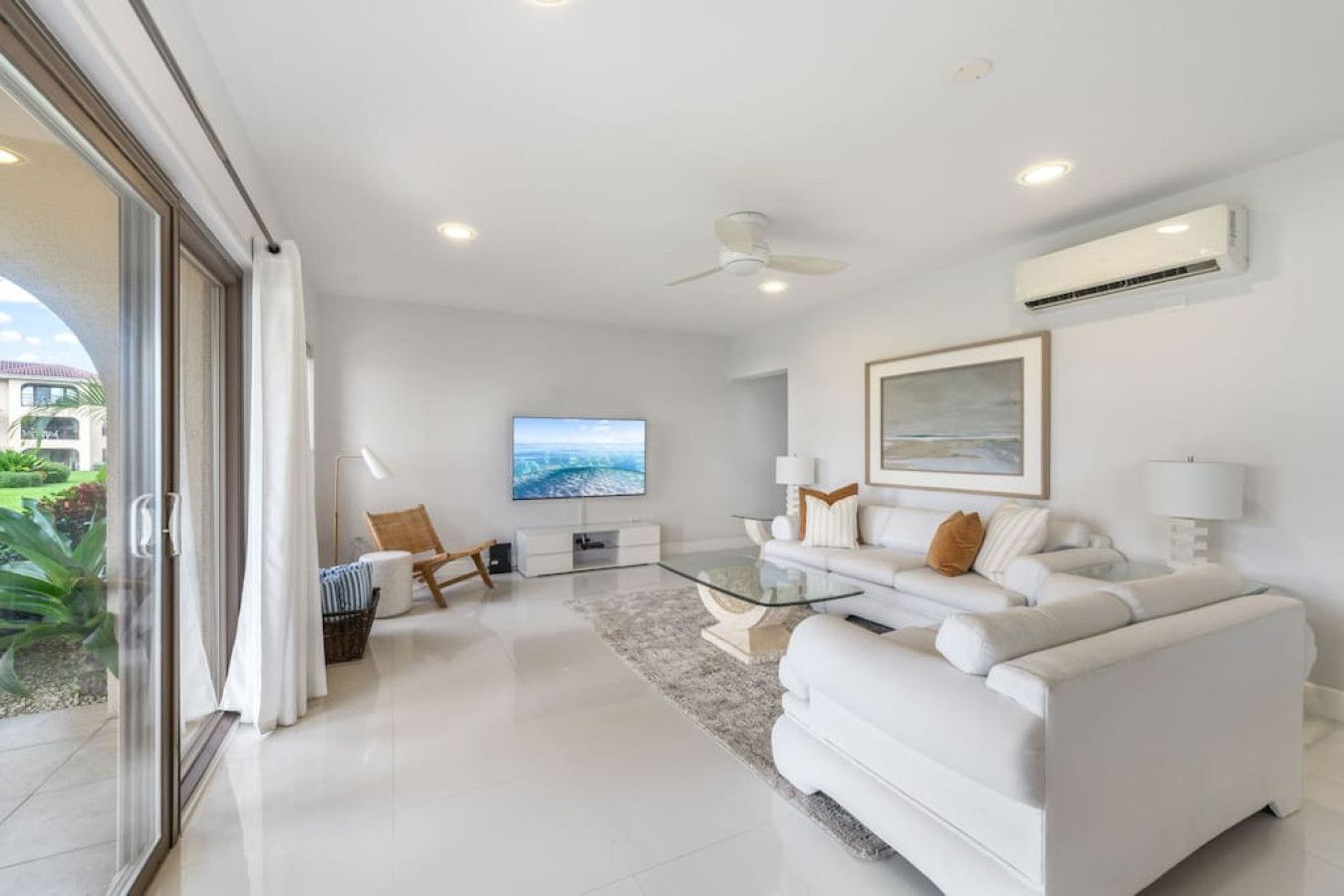 A modern living room with a glass coffee table, white sofas, a flat-screen TV, and a ceiling fan.