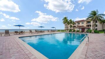 A swimming pool with lounge chairs and umbrellas, a beach view, and a building with balconies.