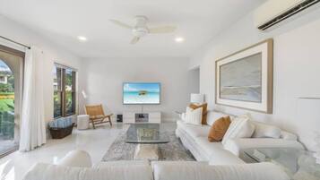 A spacious living room with a large flat-screen TV, a white sofa, a glass coffee table, and a ceiling fan.