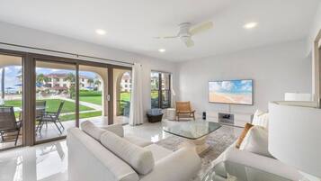 A spacious living room with a large glass coffee table, white sofas, and a flat-screen TV.