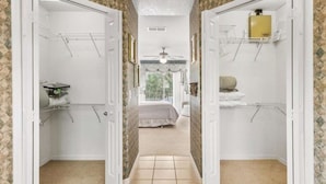 Un couloir mène à une chambre qui contient un lit, un ventilateur de plafond et offre une vue sur la verdure à l’extérieur.