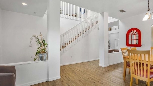 Un intérieur spacieux avec un plancher en bois, des murs blancs, un escalier, un coin repas avec des chaises en bois et une fenêtre à cadre rouge.