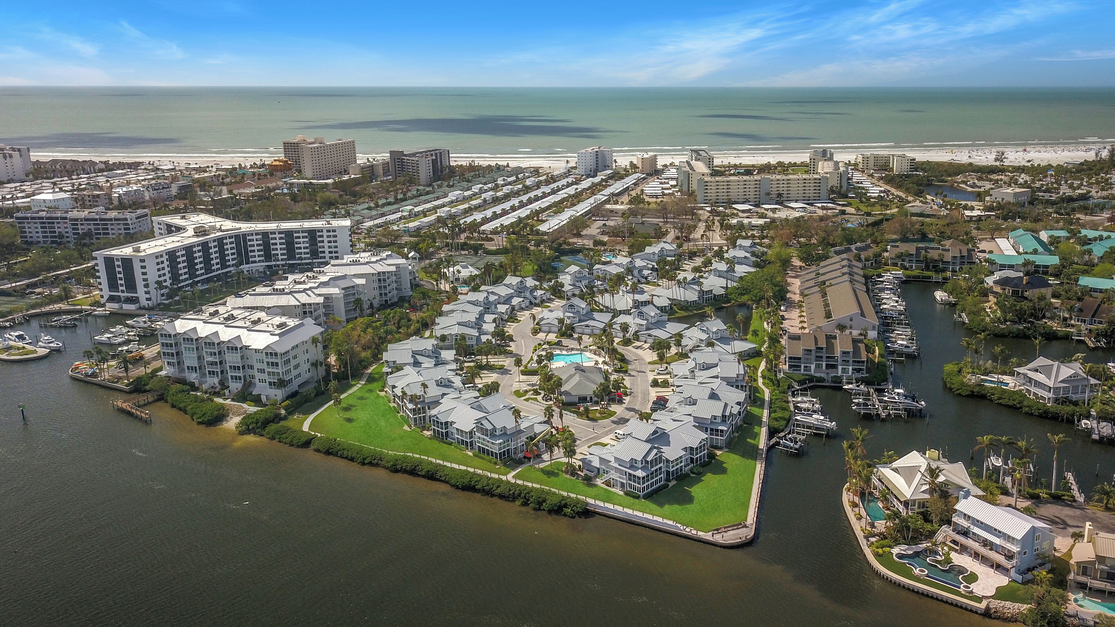 An aerial view of a coastal residential area with a marina, featuring modern multi-story buildings, a central green space, and a view of the ocean.