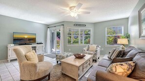A living room with a television, a ceiling fan, and a sign that reads 'BEACH HOUSE'.