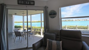 A room with a view of the beach, a sliding glass door leading to an outdoor area with a table and chairs, and a clock on the wall.