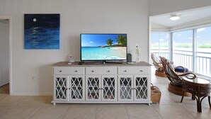 A living room with a TV displaying a beach scene, a white cabinet with glass doors, wicker chairs, and a large window offering a view of the ocean. - Bayfront Siesta Key condo with amazing views - GBB 1391 (Siesta Key)