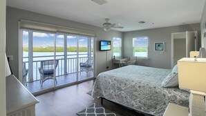 Ein Schlafzimmer mit einem großen Bett, einem Schreibtisch, einem Sessel, einem Fernseher und einem Balkon mit Sitzgelegenheiten.