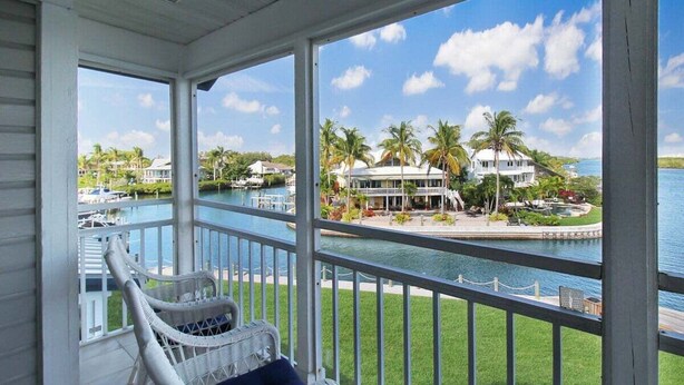 Ein überdachter Balkon mit Korbstuhl, Blick auf einen Yachthafen mit Booten und ein großes Haus mit Pool.