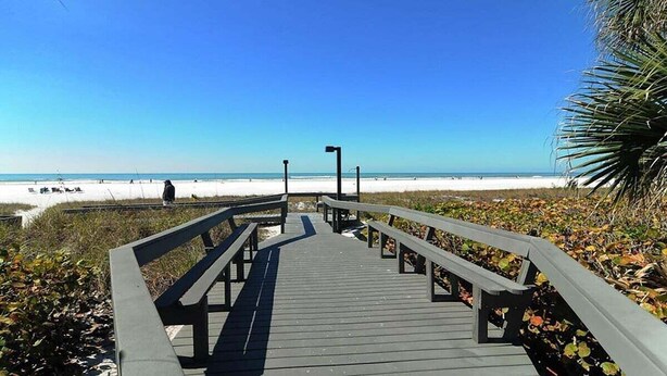 Un muelle de madera que conduce a una playa de arena blanca y un cielo azul despejado.