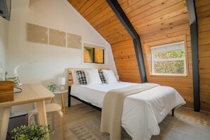 A bedroom with a bed, a desk, a chair, a window, and a ceiling with wooden beams.